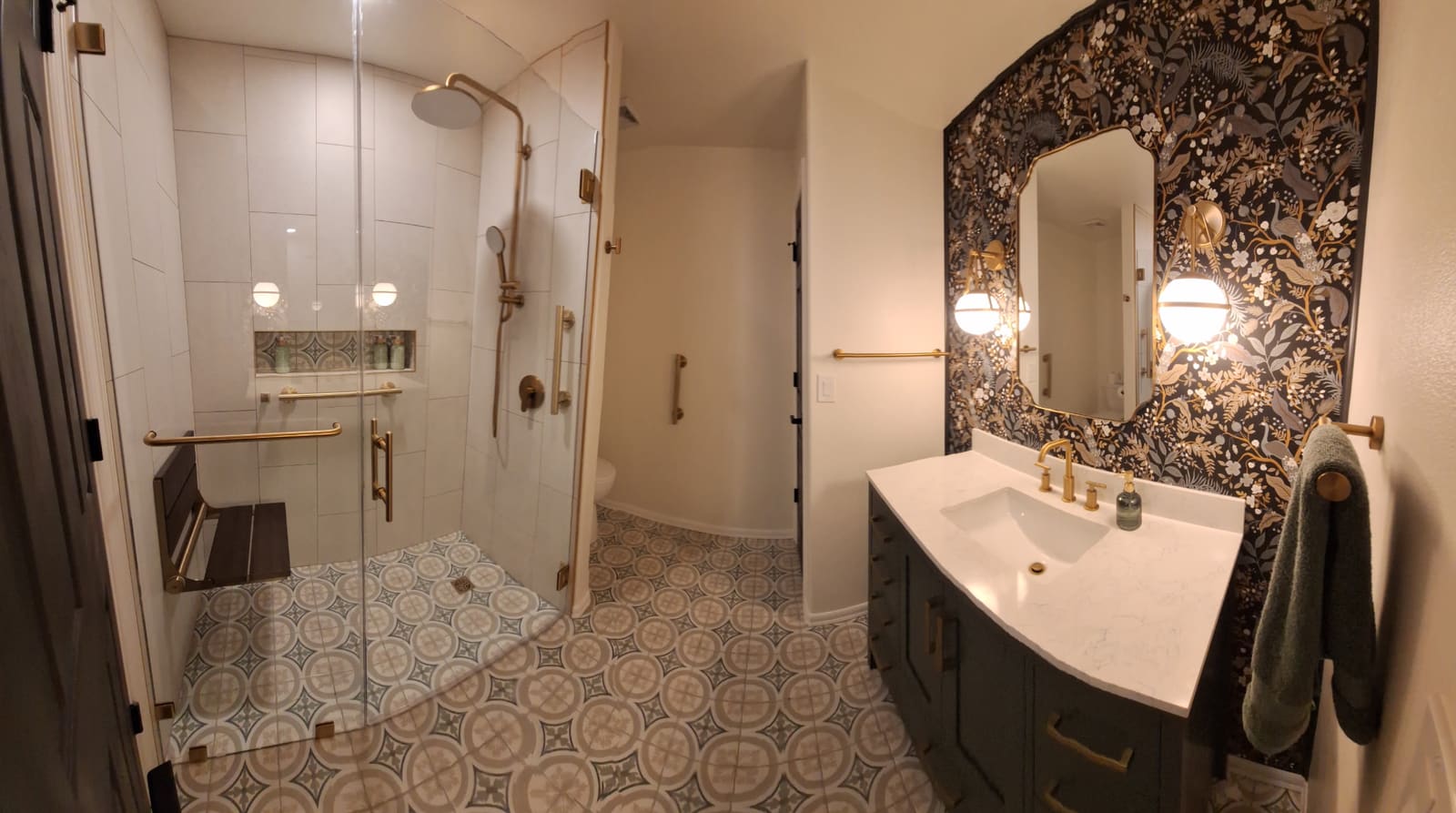 Finished bathroom with tiled walk-in shower, patterned tile floor, wallpapered vanity wall and gold fixtures